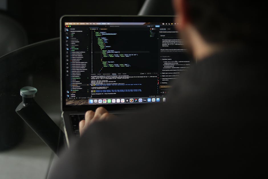 A person using a laptop with a dark screen displaying lines of colorful code, seated in a vehicle's interior. The individual’s hand is on the keyboard, and the focus is on the screen, with the person’